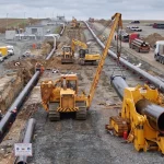 Panorama-Drohnenansicht einer Pipeline-Baustelle mit Rohrgraben, schweren Kettenbaggern, Rohrlegern und LKWs beim Verlegen großkalibriger Rohrleitungen.