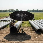 Ein großer, schwarzer Arbeitsschirm ist mittels einer Halterung direkt an einer Pipeline befestigt und spendet einem Schweißer Schatten. Der Schweißer arbeitet konzentriert an einer Rohrverbindung, während im Hintergrund zahlreiche weitere Pipeline-Rohre auf einer weiten, sonnigen Wiesenfläche lagern. Der Schirm dient hier als effektiver Sonnen- und Hitzeschutz während der Montagearbeiten im Freien.