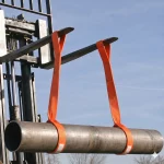 Gabelstapler hebt ein langes Stahlrohr sicher mit zwei orangefarbenen Rohrhebegurten im Außenbereich an.