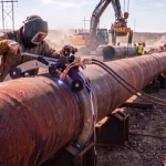 Ein Schweißer in Schutzausrüstung bedient eine manuell geführte Autogen-Rohrschneidemaschine zur präzisen Bearbeitung eines Pipeline-Rohrs auf einer weitläufigen Baustelle.