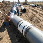 Mehrere Facharbeiter führen Schweißarbeiten an einer Pipeline durch, wobei ein Hebekissen zur stabilen Lagerung und Positionierung der schweren Rohrelemente auf der Baustelle dient.