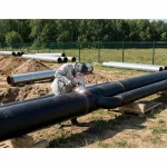 Ein Schweißer in voller Schutzausrüstung – bestehend aus einer hellgrauen Schutzjacke, Schweißerhelm und Handschuhen – arbeitet an einer schwarzen Pipeline im Freien. Während des Schweißvorgangs ist ein heller Lichtbogen mit Funkenflug an der Nahtstelle zu sehen. Die Rohre lagern auf Holzpaletten auf einem Baustellengelände, das im Hintergrund von einem grünen Zaun und Bäumen begrenzt wird.