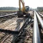 Gelber Kettenbagger auf verlegten Baggermatten aus Holz zur Bodenstabilisierung bei Pipeline-Bauarbeiten in schlammigem Gelände.