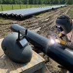 Ein Schweißer mit Schutzhelm und Arbeitshandschuhen führt Schweißarbeiten an einer schwarzen Pipeline auf einer Baustelle durch, wobei heller Funkenflug entsteht. Im Vordergrund liegt ein zweiter, baugleicher Schweißerhelm mit hochgeklapptem Visier auf einem Holzpodest. Im Hintergrund erstrecken sich mehrere parallel verlaufende Rohrleitungen über ein weitläufiges Außengelände unter sonnigem Himmel.