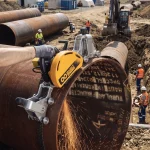 Facharbeiter trennt ein großkalibriges Stahlrohr mit einem elektrischen, kettengeführten Rohrtrennschneider unter Funkenflug auf einer Pipeline-Baustelle.